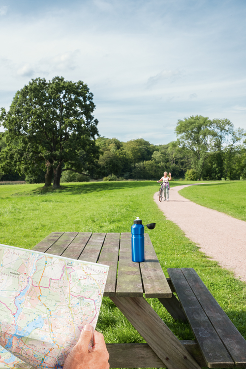 Person im Park schaut auf Landkarte (Nahbereich), Getränkeflasche (mittlerer Bereich) und erblickt eine Person mit Fahrrad im Fernbereich